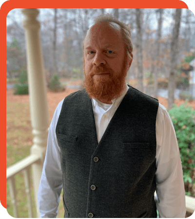 A man with red beard and white shirt standing on porch.